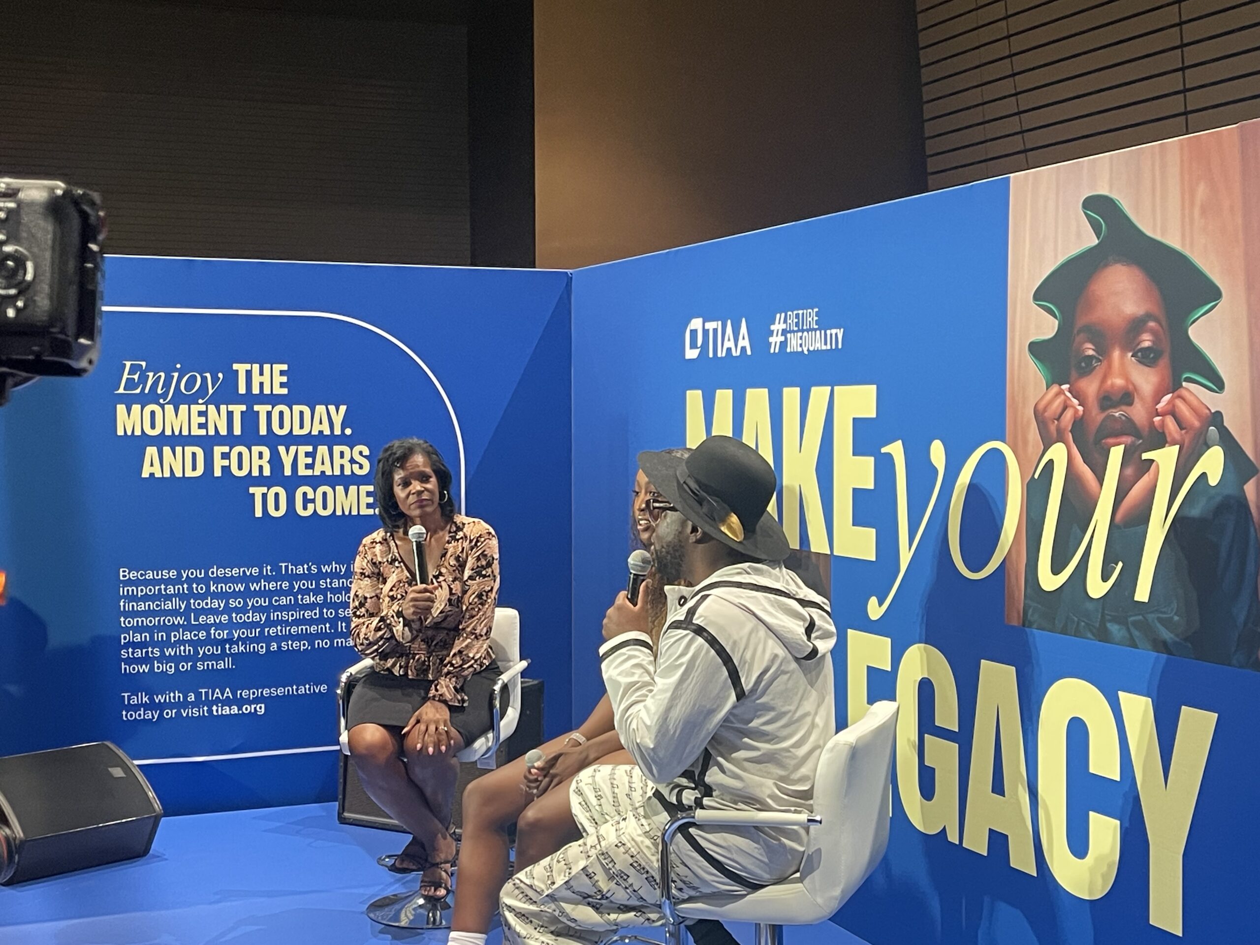 Three people sit and talk on a stage with TIAA branding and a large “Make Your Legacy” sign, next to an image of a woman and an informational display about financial planning as part of TIAA’s Financial Education Campaigns.