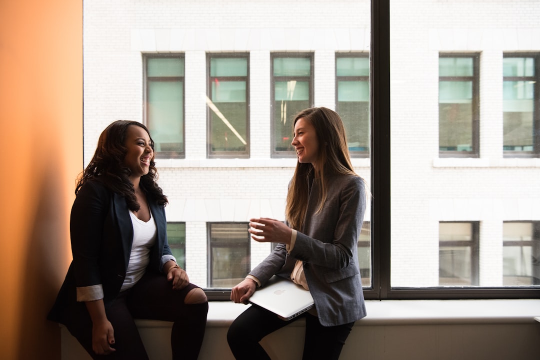 Two women in business attire sit on a windowsill, engaged in conversation about fintech partnerships, as one holds a closed laptop. Large windows behind them reveal another building.