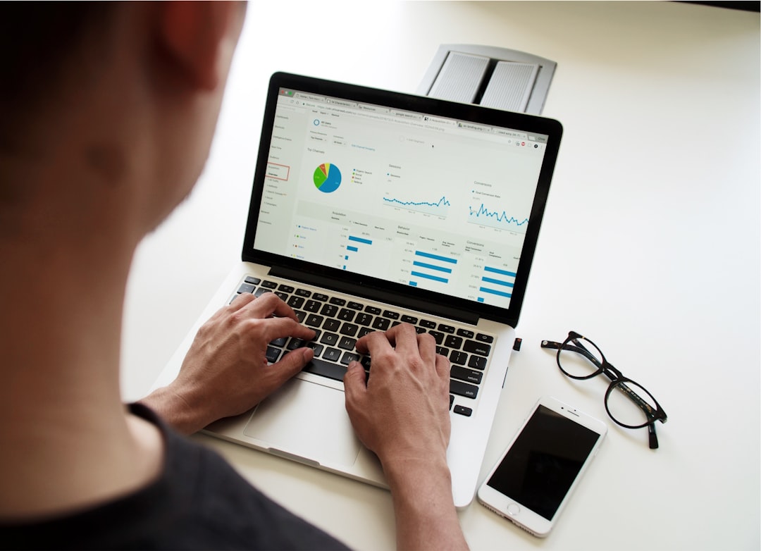 A person intently types on a laptop displaying charts and graphs, delving into the fascinating world of emotional finance. A smartphone, glasses, and a pen are neatly arranged on the desk, ready for any insights or analyses that might arise.