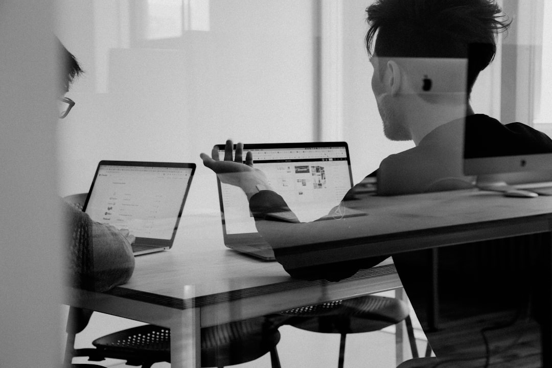 Two people working on laptops at a table, viewed through a glass partition. One appears to be explaining something related to financial partnerships, gesturing toward the screen as they collaborate effectively.