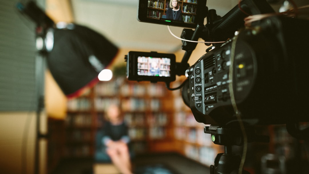 A video camera focuses on a person seated in a library, surrounded by monitors displaying the scene. Studio lights enhance the professional setup, creating an ideal environment for discussing financial journalism.