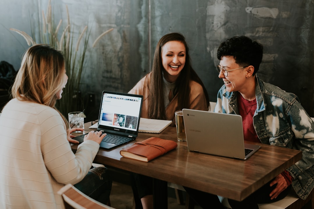 Three people sit at a table, working on laptops and smiling, embodying the spirit of education partnerships, with a notebook and glass adding to the collaborative ambiance.