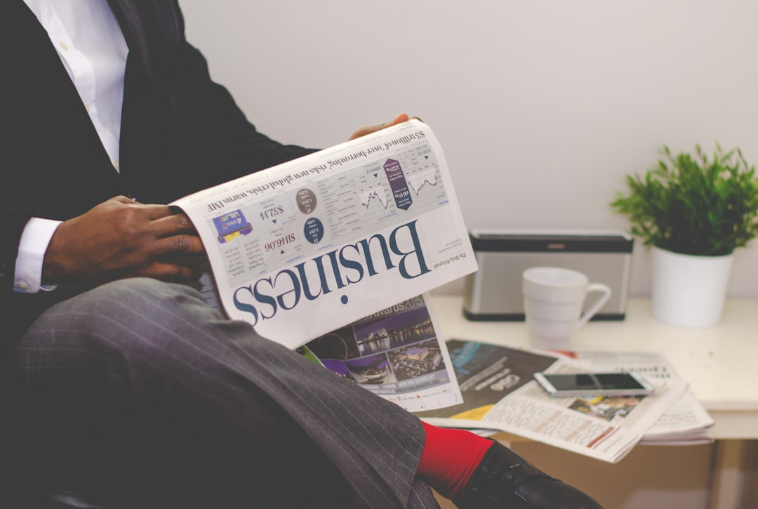 A person in a suit reads a business newspaper on financial inclusion while sitting near a table with a plant, white mug, and smartphone in a minimalist office setting.