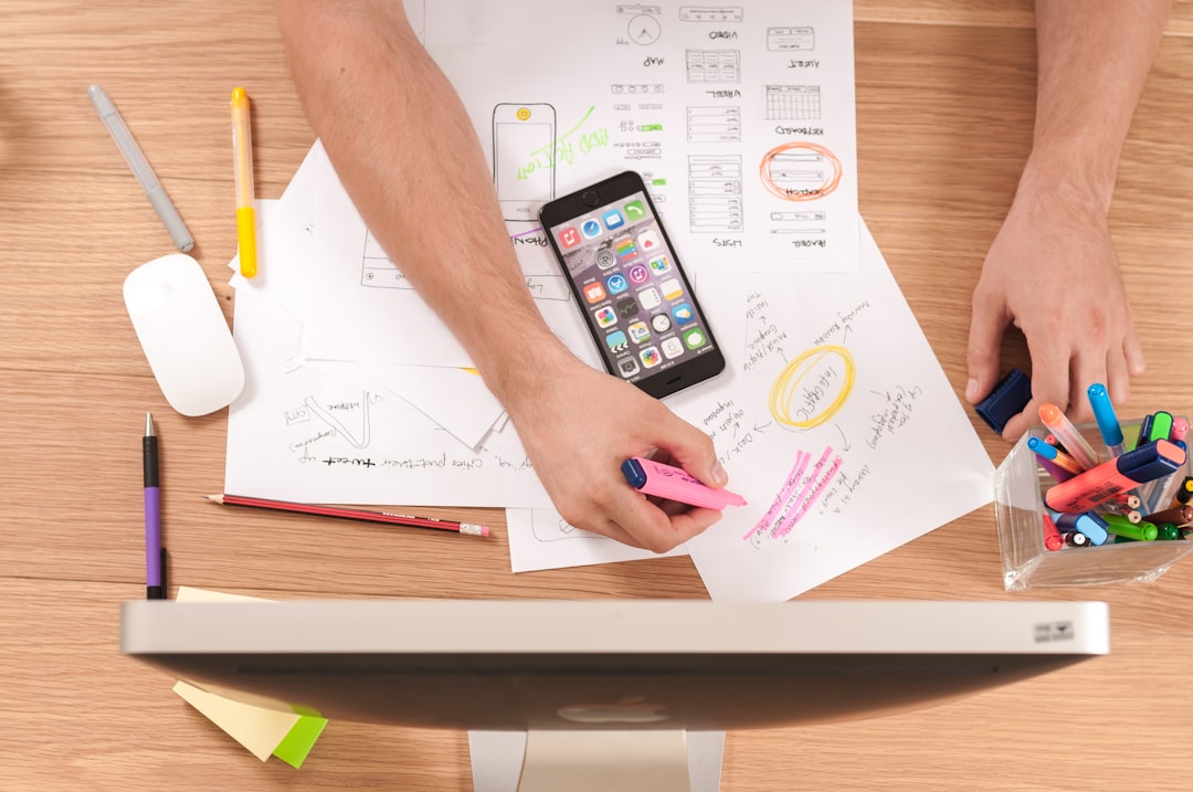 A person is diligently working at a desk, surrounded by a computer, smartphone, and papers. They highlight notes with a pink highlighter amidst various pens and pencils, focused on advancing their financial planning education.
