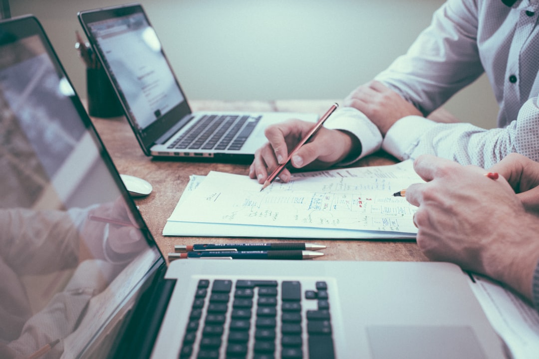 Two people collaborate at a wooden table with two laptops and scattered papers, using a pencil to discuss notes and plans, focusing on maximizing education ROI.