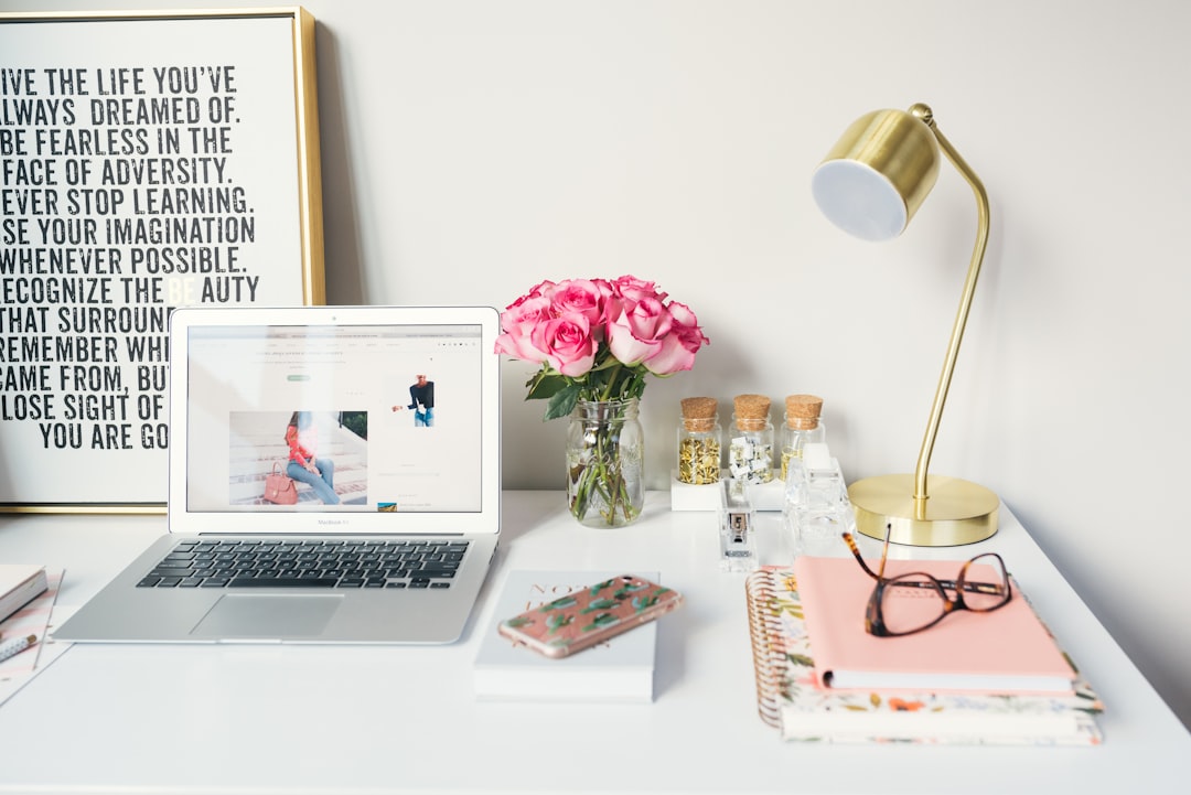 A tidy desk features a laptop showcasing an AI financial education website, pink roses in a vase, a gold lamp, a phone, notebooks, glasses, and small jars. A framed motivational quote leans against the wall.