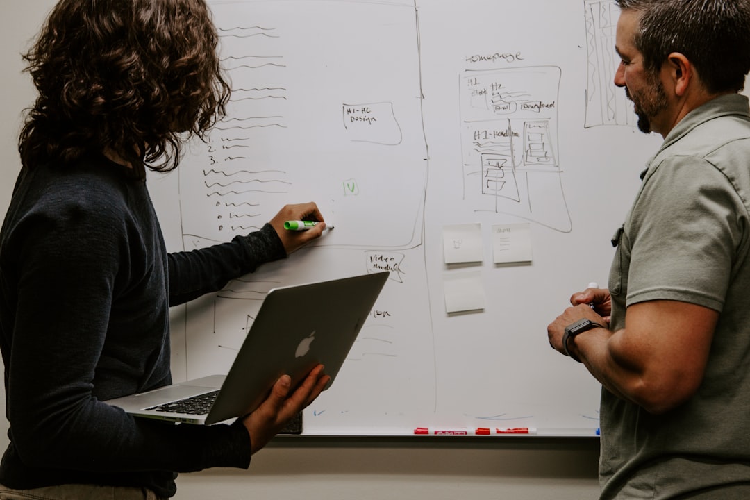 Two people discuss at a whiteboard filled with diagrams and notes focused on financial literacy. One holds a laptop and writes with a marker, while the other listens attentively.