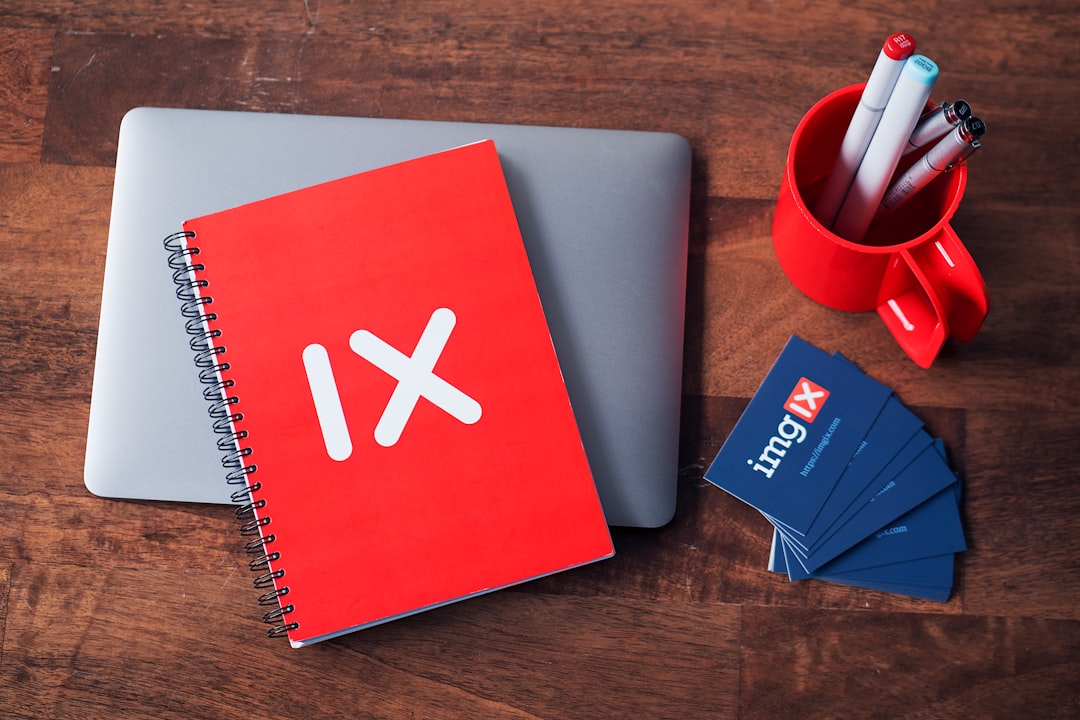 Red notebook labeled "IX" rests on a closed laptop, surrounded by blue business cards hinting at financial networking, while a red mug full of pens adds a pop of color to the wooden table's surface.