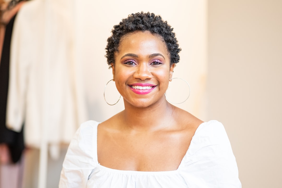 Smiling person with short curly hair in a white blouse, wearing hoop earrings, stands in a softly lit room with blurred background.