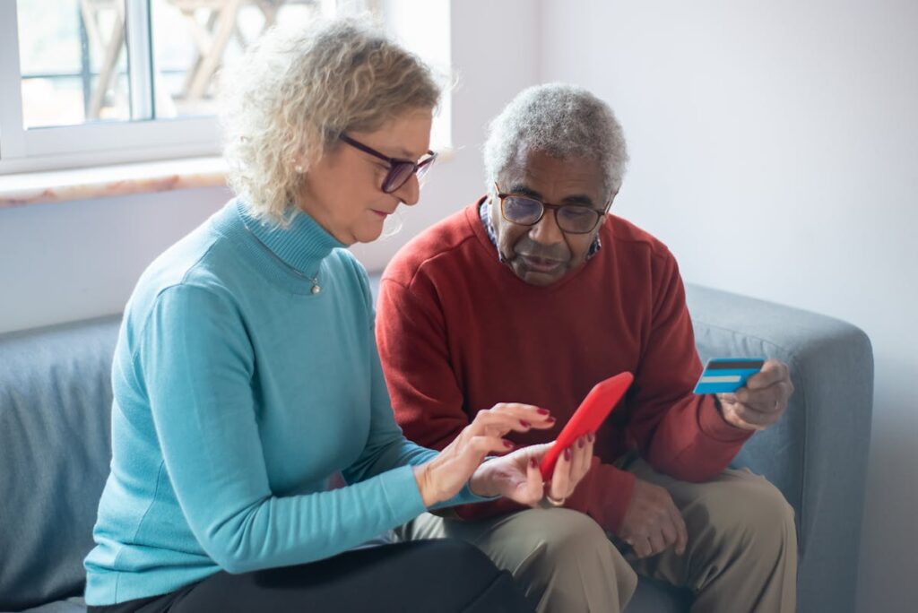 Two older adults sit on a couch; one holds a smartphone and the other a credit card, both looking at the phone screen as they explore financial education for seniors.