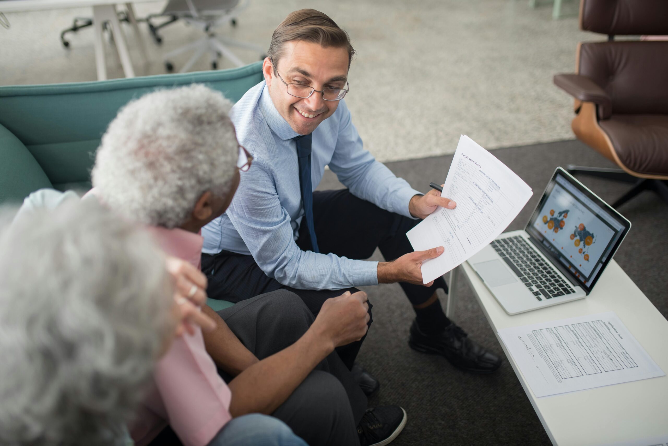 A man in business attire shows documents to two older adults while sitting by a laptop displaying a map on a screen in an office setting. Financial Education for Seniors