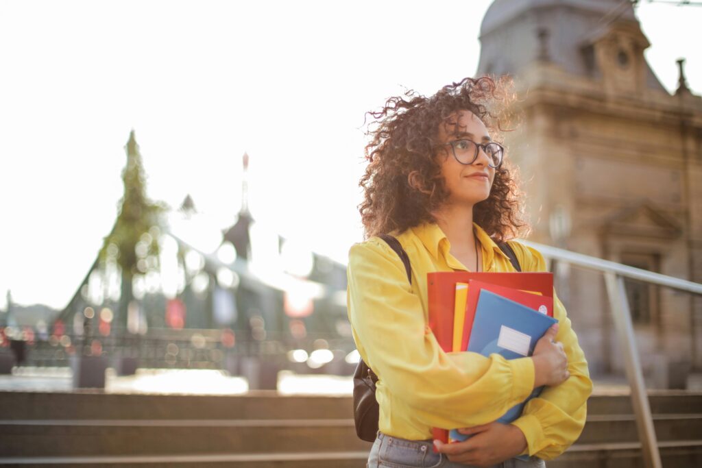 women and financial wellness: A young woman with curly hair and glasses, wearing a yellow shirt, stands outdoors holding notebooks and folders, with a historic building in the background.