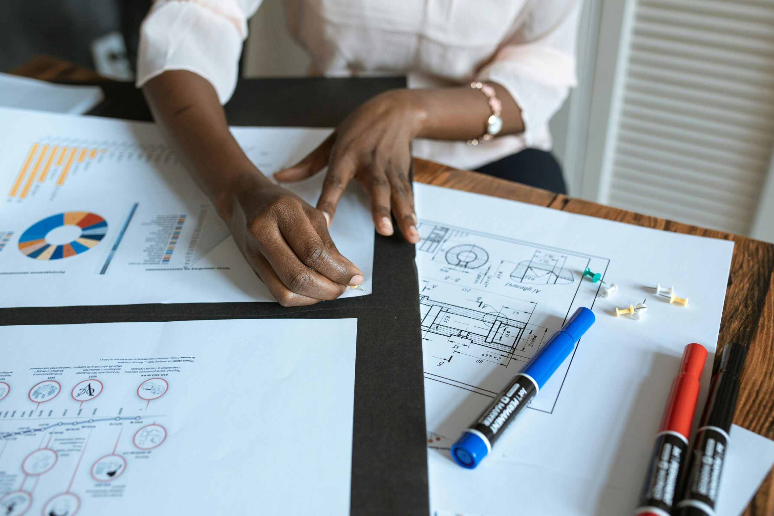 Person working at a desk with charts, graphs, and architectural plans, using a pencil. Blue, red, and black markers are also on the table. women's financial survey