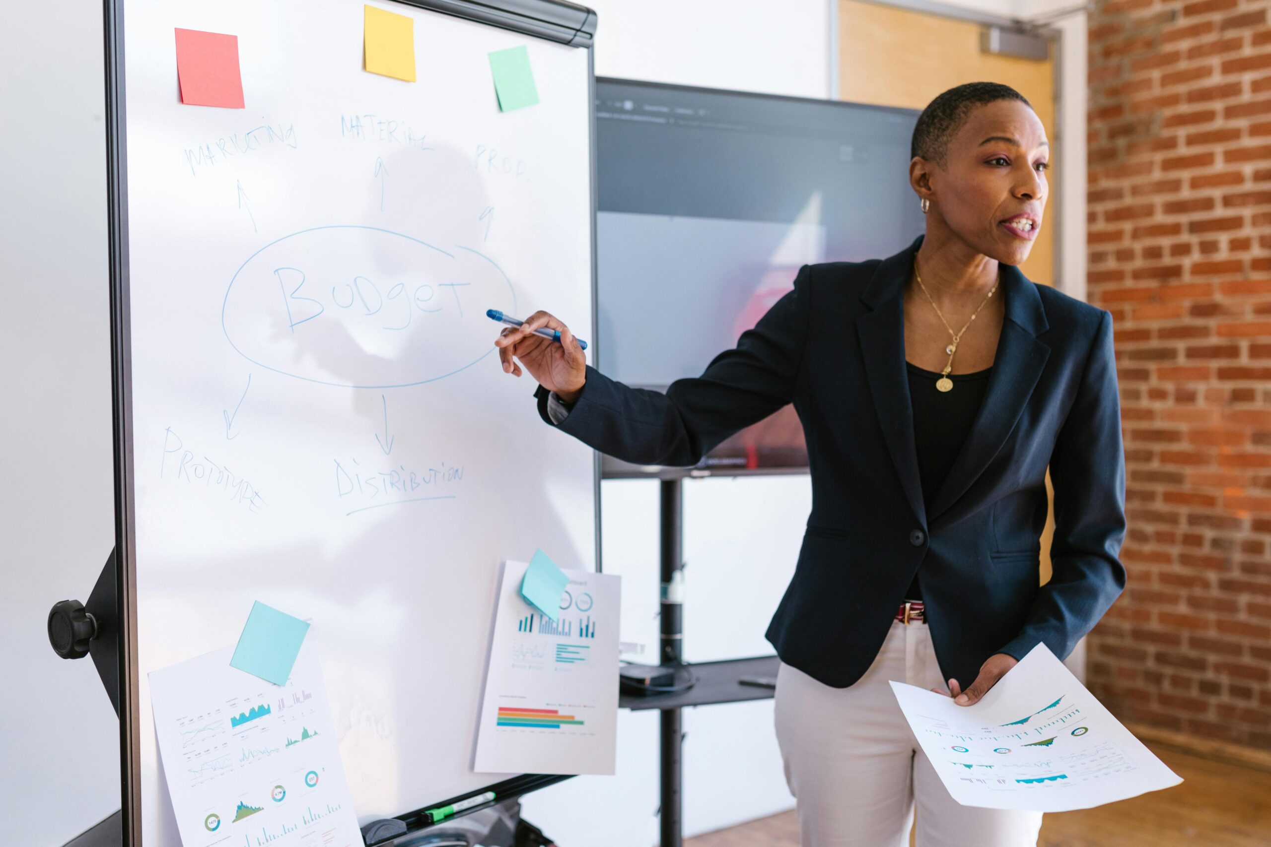 A person in business attire stands by a whiteboard labeled "Budget," holding papers and gesturing while presenting financial information. financial education training