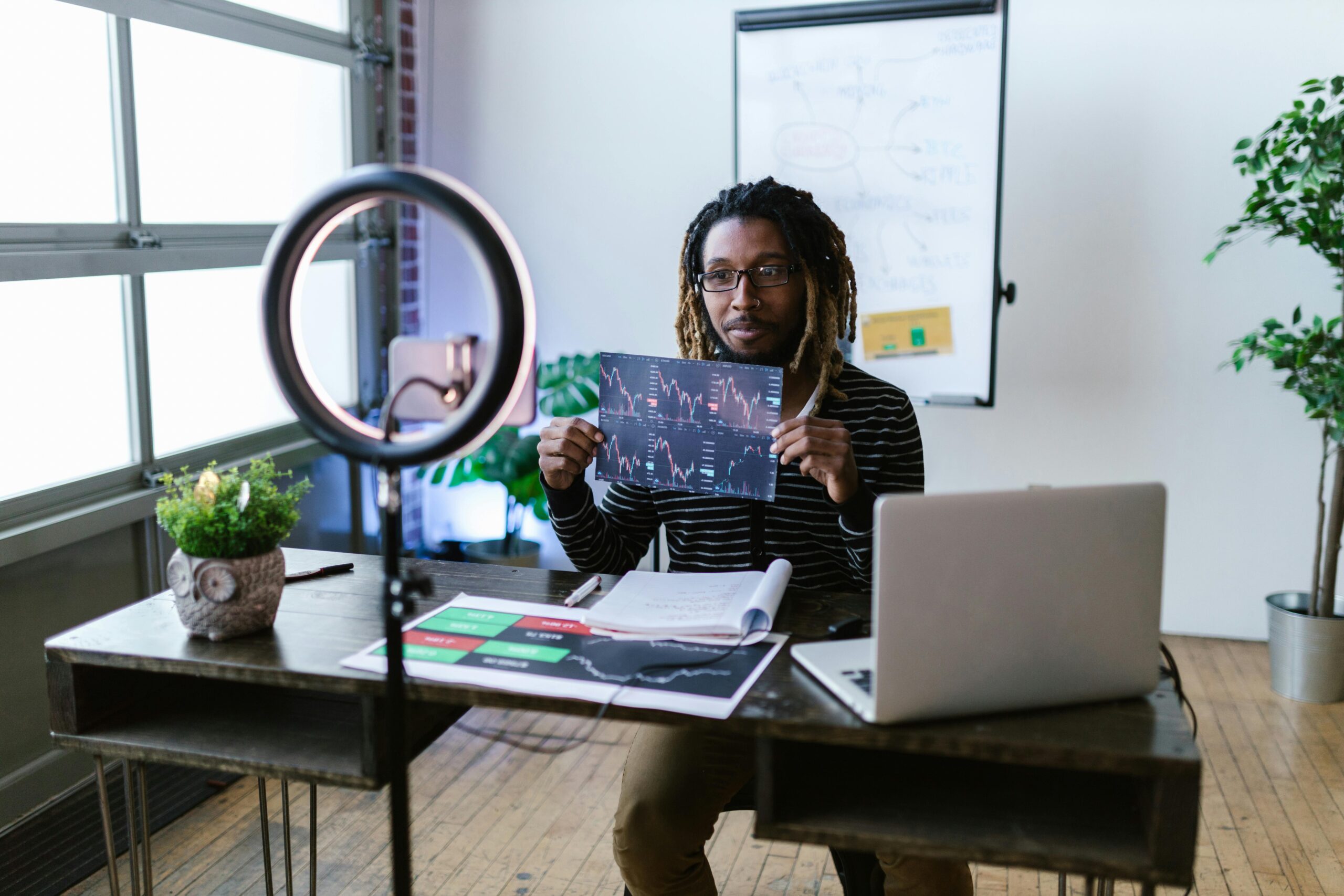A person sits at a desk with a laptop, holding up a chart in front of a ring light, with a whiteboard and plants in the background. financial education content