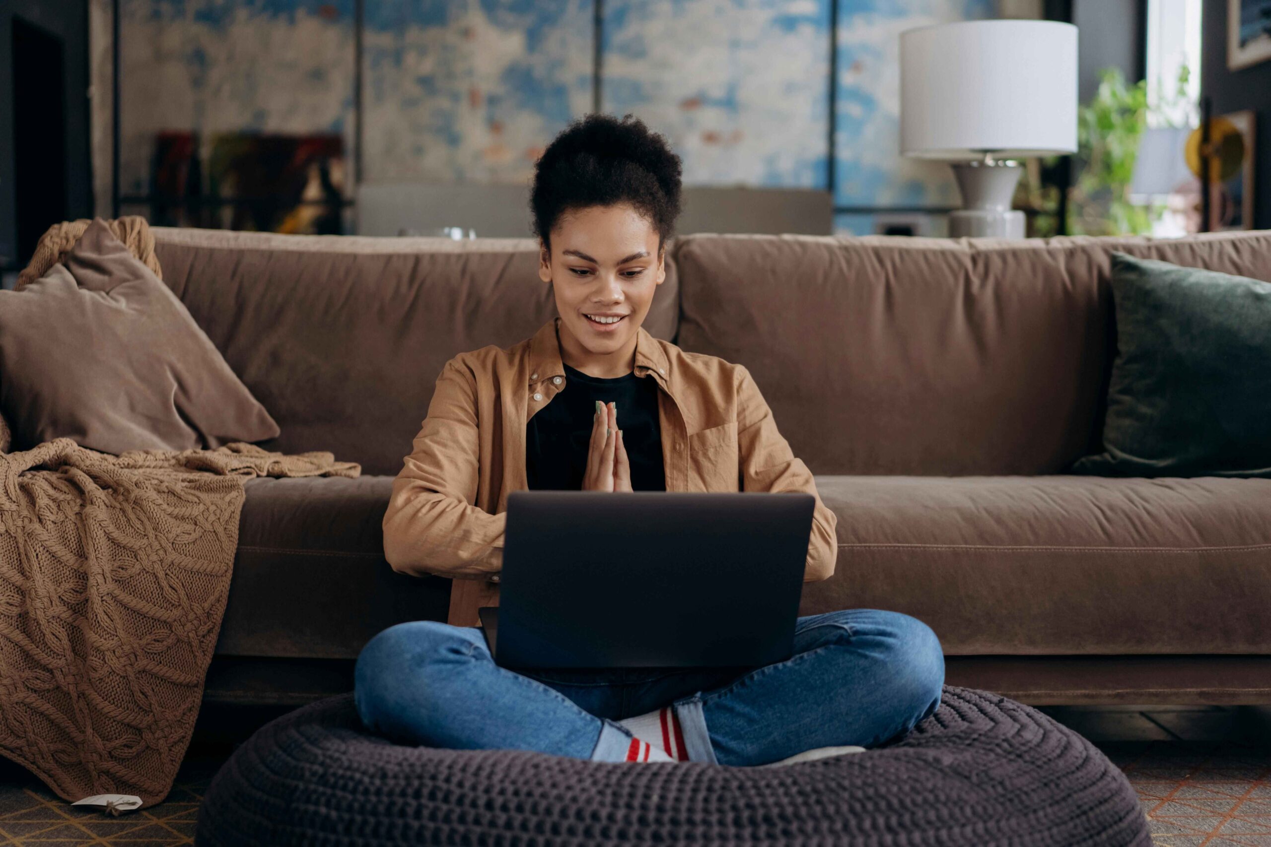 A person sits cross-legged on a cushion in front of a laptop, hands pressed together in a gesture, with a sofa and lamp in the background. women's financial literacy