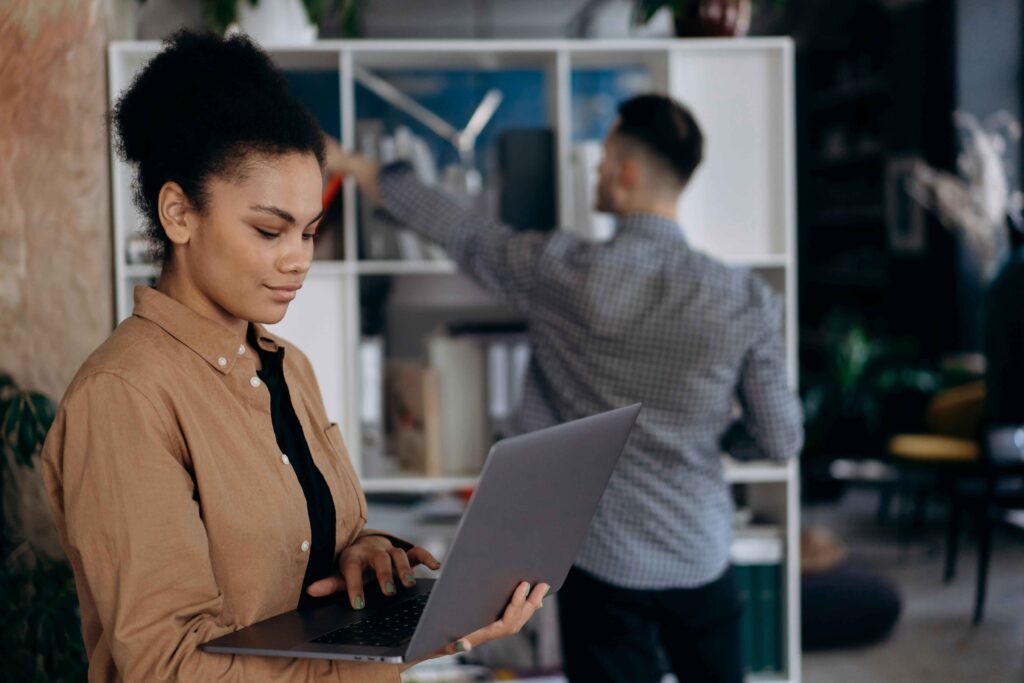 A woman stands holding a laptop and typing while a man in the background reaches for something on a shelf in an office setting. debt free movement