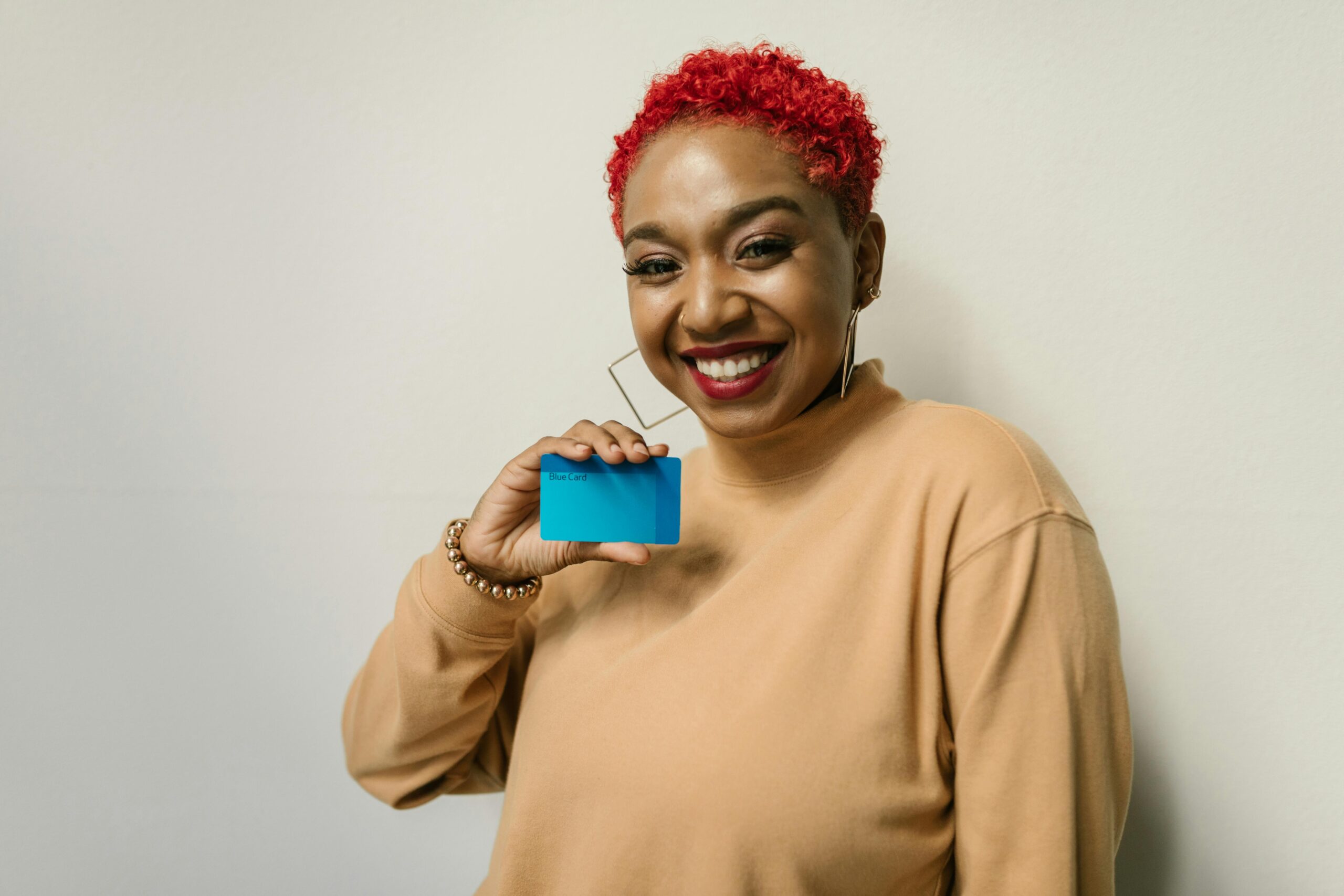 A woman with short red hair and large earrings smiles and holds up a blue card against a plain light-colored background. savings campaign