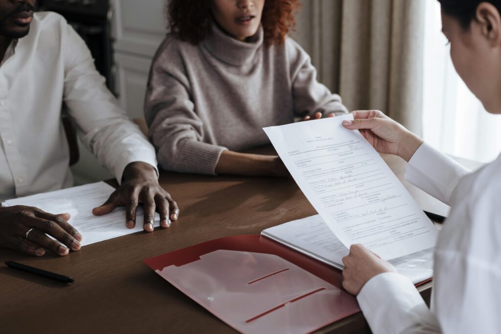 Three people sit at a table with documents; one person is holding and reviewing a paper form while the others listen. black women and personal finance