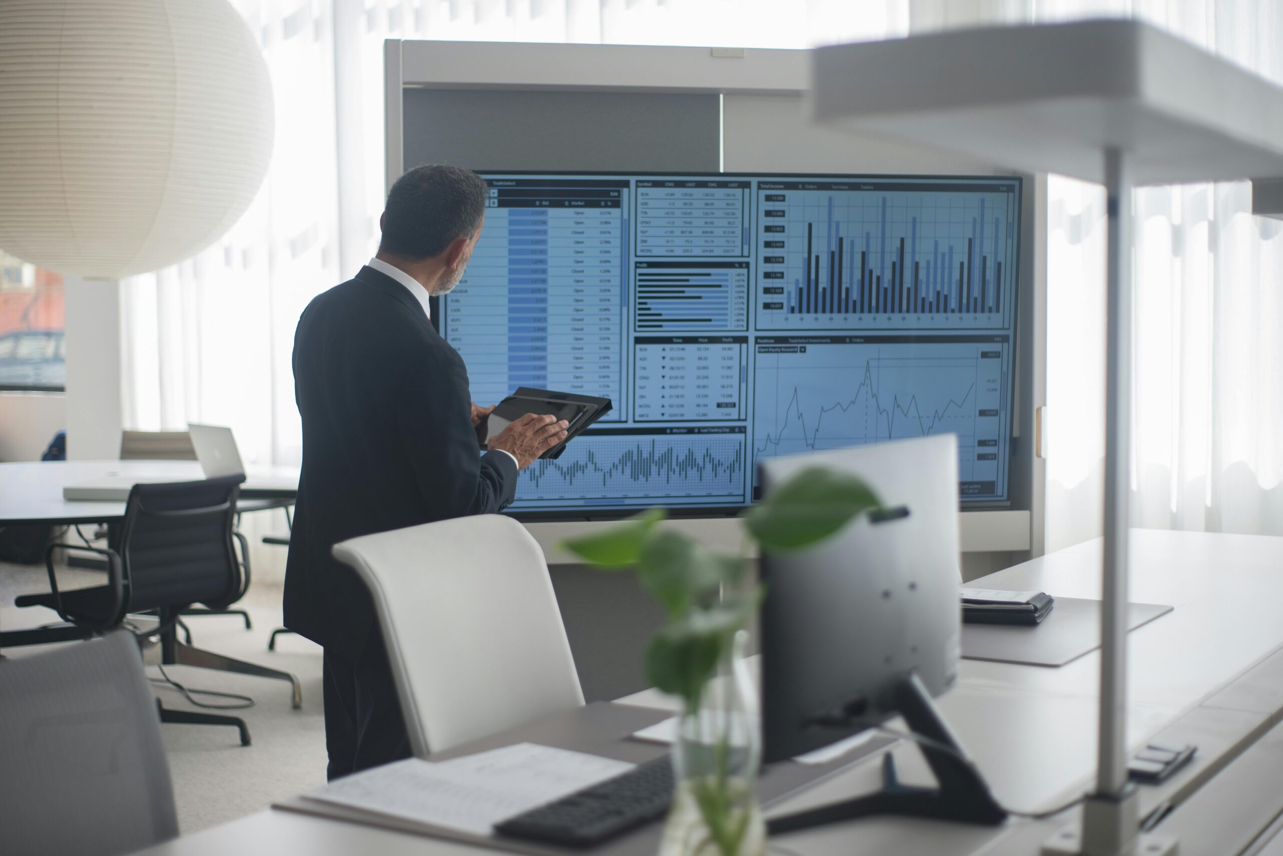 A man in a suit views financial charts and data on large monitors in a modern office, holding a tablet. Financial integrity