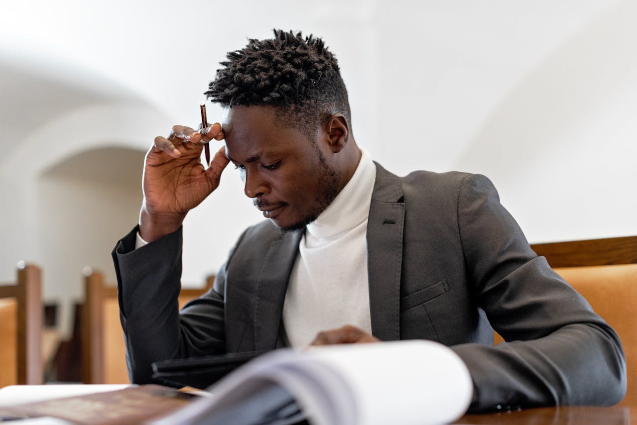 A man in a suit sits at a table, holding a pen to his head and reading documents, appearing focused and thoughtful as he contemplates corporate social responsibility.