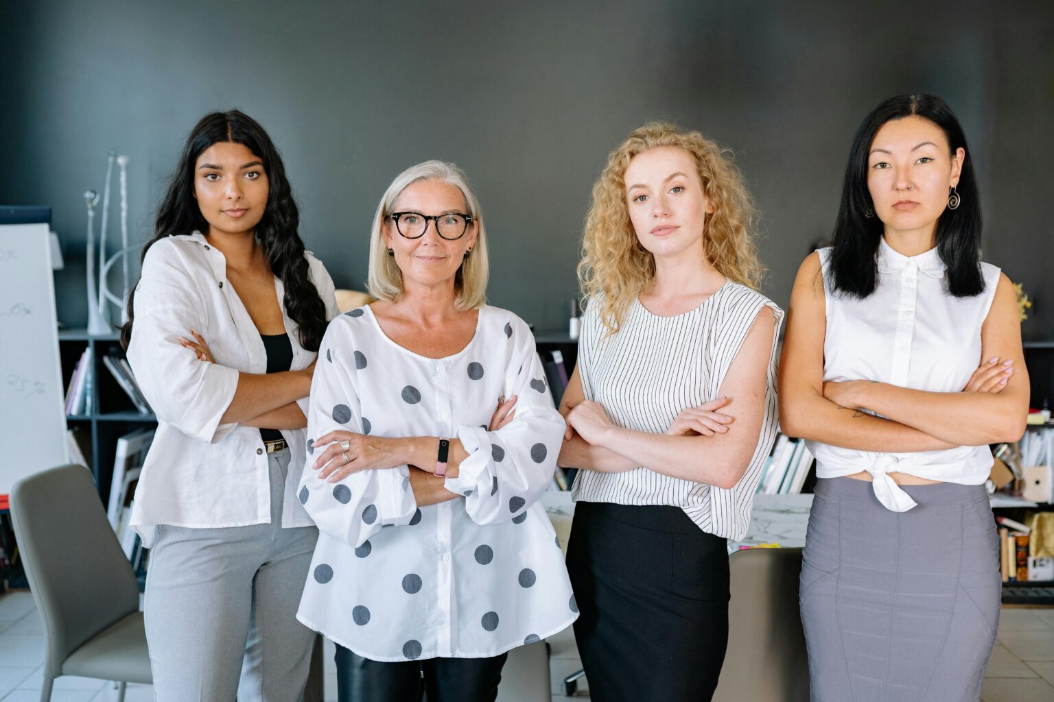 Four women stand side by side in a modern office, all facing the camera with their arms crossed, dressed in professional attire. corporate sponsorships for influencers