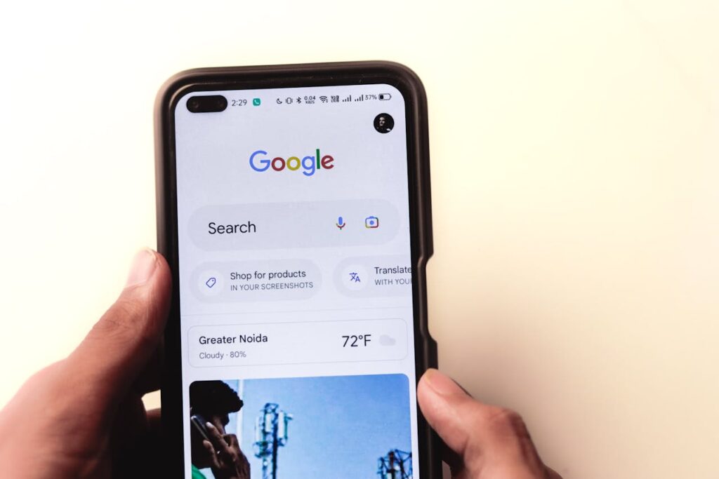 A person holds a smartphone displaying the Google search homepage, showing search options, weather, and other features on the screen.